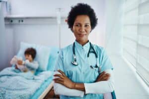 Black children's nurse with her arms crossed at hospital ward looking at camera.