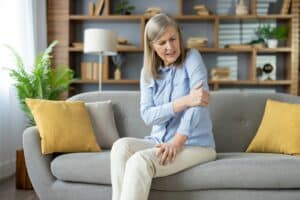 Mature woman having elbow injury and massaging sore spot with hand in stylish apartment interior