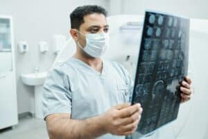 Serious male radiologist in mask and uniform looking at x-ray image of patient